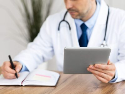 Healthcare, Medical Care And Technology Concept. Male doctor working in office at clinic, writing with pen in notebook, holding and using digital tablet, checking appointment schedule